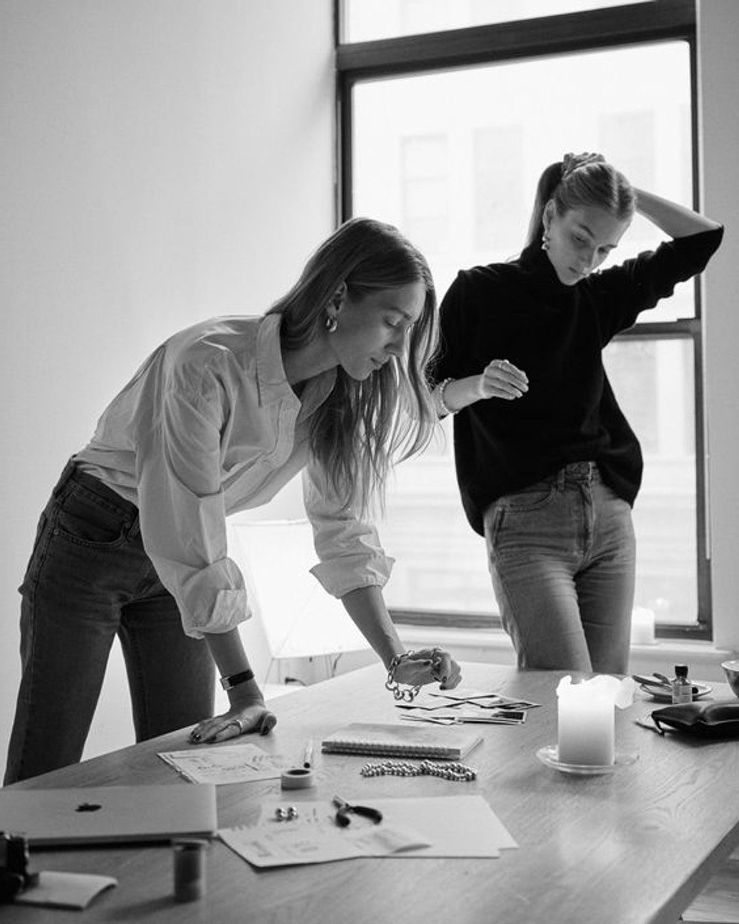 architectural-review Two individuals in casual attire work at a table in a well-lit room. One leans over documents while the other stands, hand on head. A lit candle and various items are on the table.