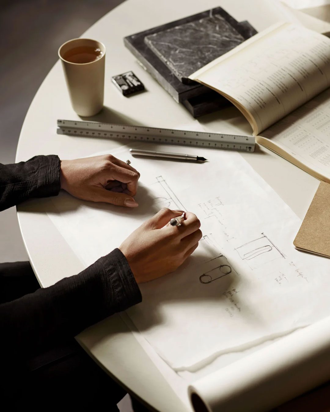 furnishes Person drawing technical sketches on a table with a ruler, book, eraser, and coffee cup.