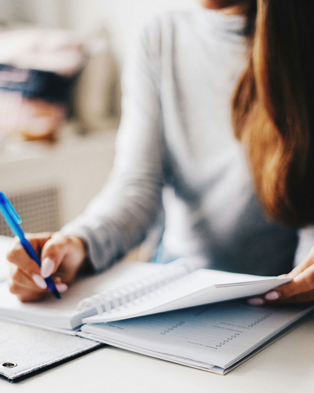 procurement A person wearing a grey sweater writes in a notebook with a blue pen while flipping through its pages.