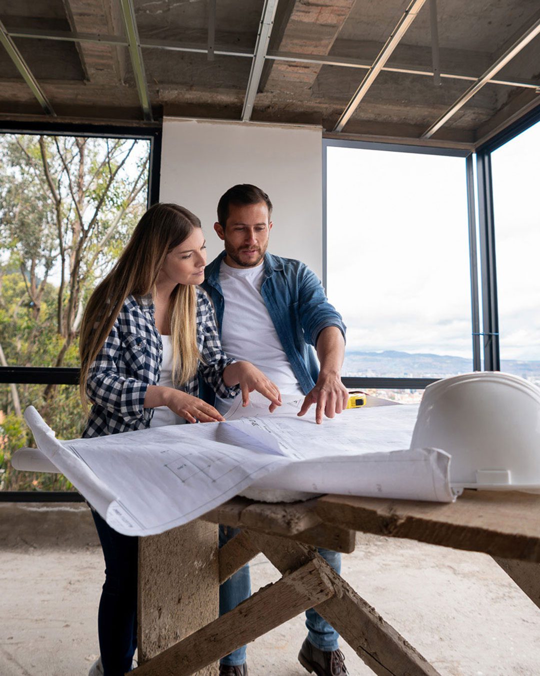 project-management Two people standing in a partly constructed building, looking at a large blueprint on a wooden table with a white hard hat placed nearby.