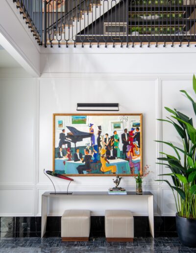 A two-story room with a staircase, modern painting, two beige ottomans, side table with plants, and sculpture. Large plant on the right.