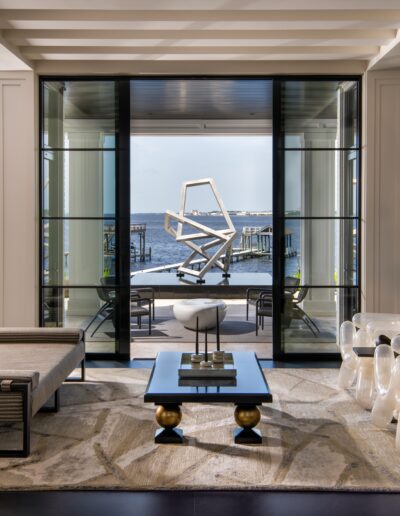 A modern living room with large glass doors opening to a view of a waterfront deck. There is contemporary furniture, including a lounge chair, a coffee table, and a grand piano. A metal sculpture is outside.