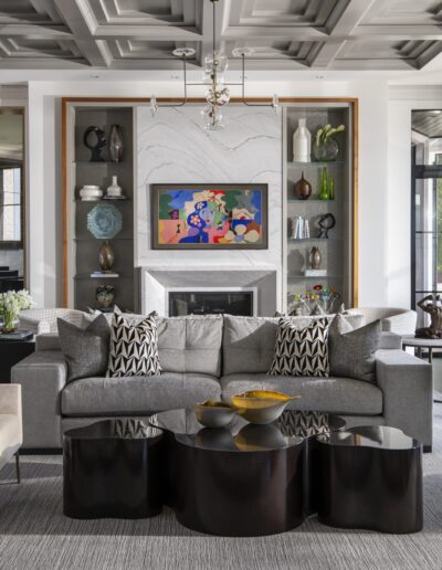 Modern living room with patterned ceiling, gray sectional sofa, black coffee tables, colorful artwork above the fireplace, and large windows letting in natural light. A piano is on the right side.