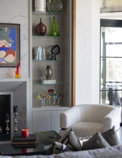 A living room with a modern aesthetic featuring a grey chair, glass coffee table, abstract art, and a built-in shelf displaying decorative items like vases, sculptures, and a colorful flower arrangement.