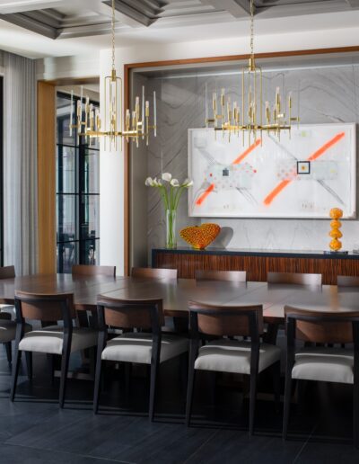 A modern dining room with a large wooden table, twelve chairs, two chandeliers, wall art, and a sideboard with decorative items. Large windows provide natural light.