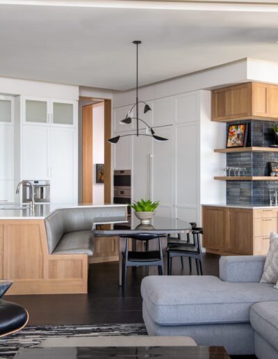 Modern kitchen and living space with white cabinetry, light wood accents, an L-shaped gray sofa, and a contemporary black pendant light fixture. Large windows illuminate the area with natural light.