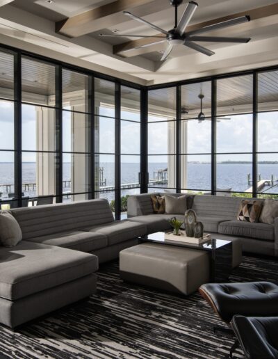 Modern living room with large windows offering a view of a lake. Room features a sectional sofa, two ottomans, a patterned rug, and ceiling fans. Outdoor seating area visible.