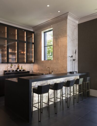 A modern home bar with a marble countertop, black stools, a sleek sink, and backlit shelving with bottles, illuminated by recessed lighting.