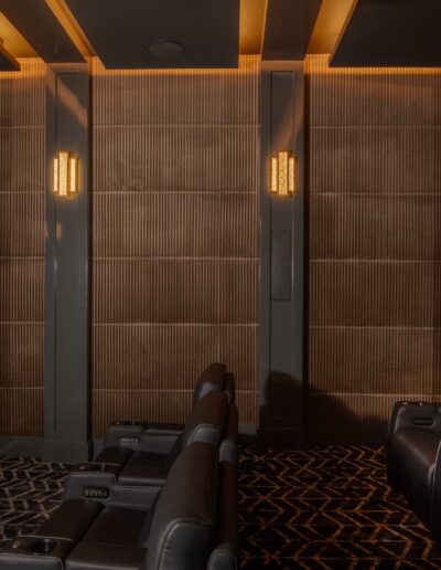 A dimly lit, modern home theater with black recliner seats arranged in rows, illuminated wall sconces, shaded panels, and a patterned carpet.