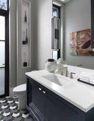 A modern bathroom with a patterned black-and-white floor, dark vanity with white countertop, white sink, towel, vase, wall decor, large mirror, and frosted glass door.