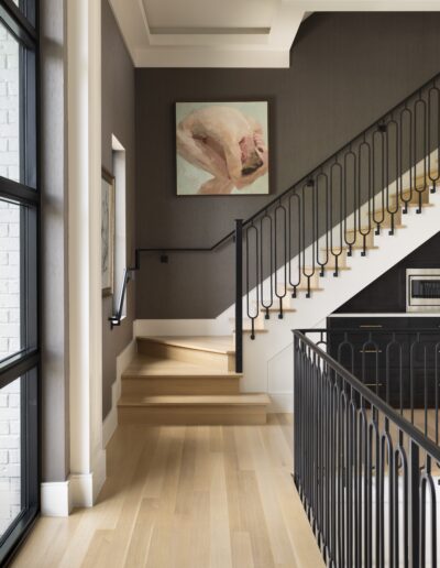 Modern staircase with light wood steps and black railing in a well-lit interior. A large abstract painting hangs above the stairs, and large windows provide ample natural light.