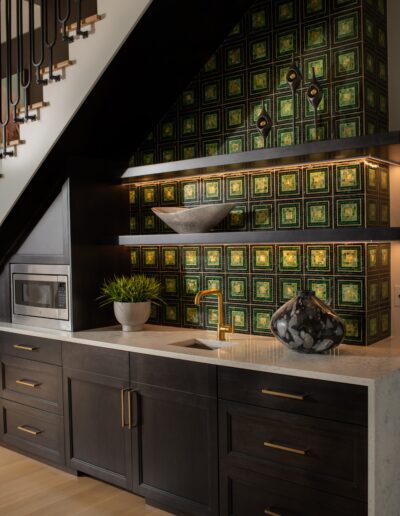 A modern kitchen nook featuring dark cabinetry, a sink, a microwave, and decorative green tiled backsplash under a staircase.
