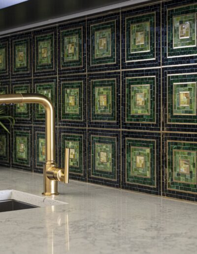 A kitchen countertop with a brass faucet, a small sink, a potted plant, and a green and black tiled backsplash.