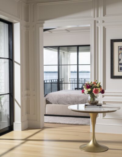A light-filled room with glass doors leading to a balcony overlooking water. A round table with a floral arrangement stands in the foreground, and a bed is visible in the adjacent room.