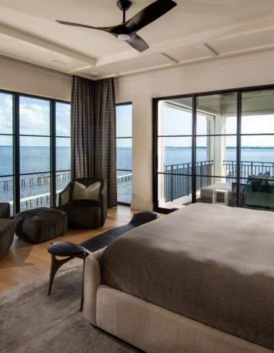 A bedroom with large windows offers a scenic water view. The room features a bed, seating area, and contemporary decor. A balcony with outdoor furniture is accessible through glass doors.