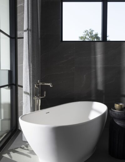 A modern bathroom with a freestanding white bathtub under a rectangular window, black side table with a candle, and floor-to-ceiling glass door with curtains.